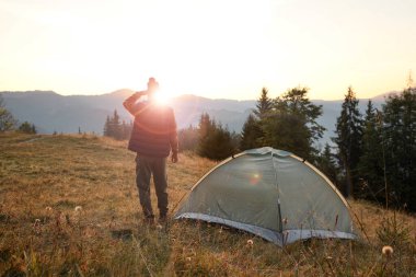 Gün batımında dağlarda kamp çadırının yanında bir adam var.