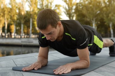 Sportif adam nehir kıyısındaki minder üzerinde kalas egzersizi yapıyor.