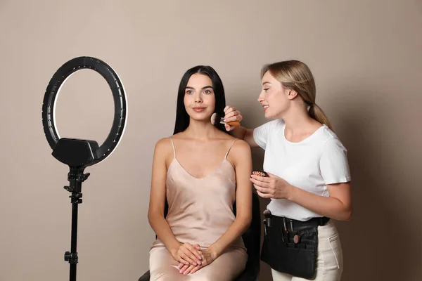 ベージュを背景に美しい若い女性と働くプロの化粧アーティスト リングランプの使用 — ストック写真