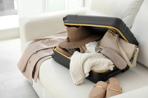 Open suitcase full of clothes, jacket and fashionable shoes on sofa in room