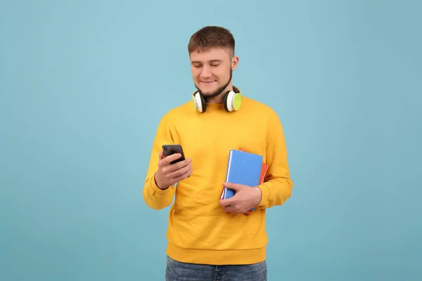 Jeune Étudiant Avec Écouteurs Livres Utilisant Smartphone Sur Fond Bleu — Photo