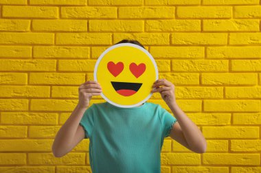 Sarı tuğla duvarın yanında kalbi olan küçük bir kız emojisi.