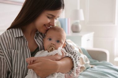 Mutlu anne küçük bebeğini kucağında emzik ile evde tutuyor, mesaj atmak için yer var.