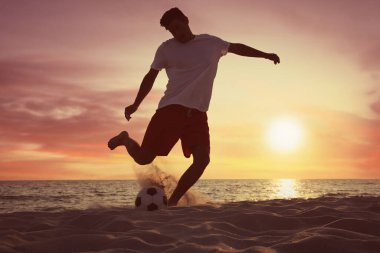 Adam gün batımında plajda futbol oynuyor.