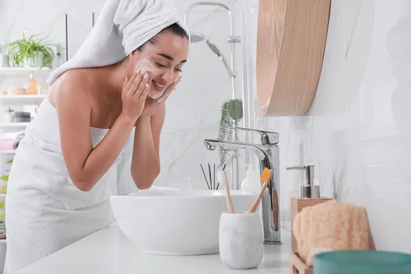 Beautiful young woman applying cleansing foam onto face in bathroom. Skin care cosmetic