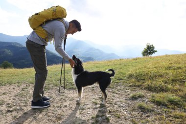 Sırt çantalı ve sırıkla gezen bir turist dağlarda sevimli bir köpeği okşuyor.