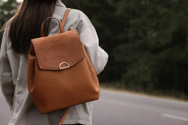 Young woman with stylish backpack near empty road in forest, back view. Space for text