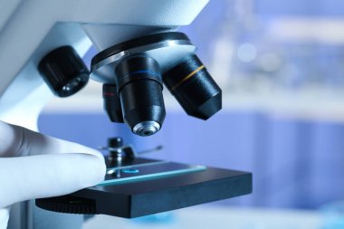 Bilim adamı laboratuarda mikroskop altında açık mavi sıvı örneğini inceliyor.