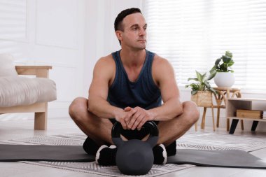Evde kettlebell 'li kaslı bir adam oturuyor.