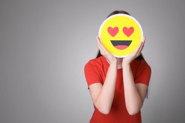 Woman covering face with heart eyes emoji on grey background, space for text clipart