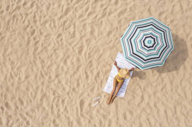 Kumlu sahilde çizgili plaj şemsiyesi altında dinlenen bir kadın, hava manzaralı. Metin için boşluk