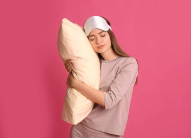 Pembe arka planda, uyurgezer halde yastıklı pijama ve maske giyen genç bir kadın.