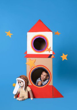 Açık mavi arka planda karton roketle oynayan sevimli küçük çocuk.