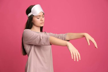 Pembe arka planda uyurgezer halde pijama ve maske giyen genç bir kadın.
