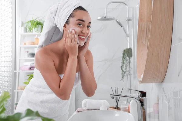 在浴室靠近镜子的地方 漂亮的年轻女子在脸上涂上清洁剂泡沫 护肤化妆品 — 图库照片