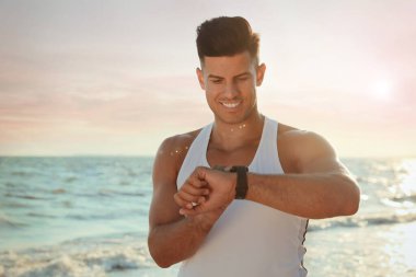 Spor vücutlu bir adam plajda spor bilekliğini kontrol ediyor.
