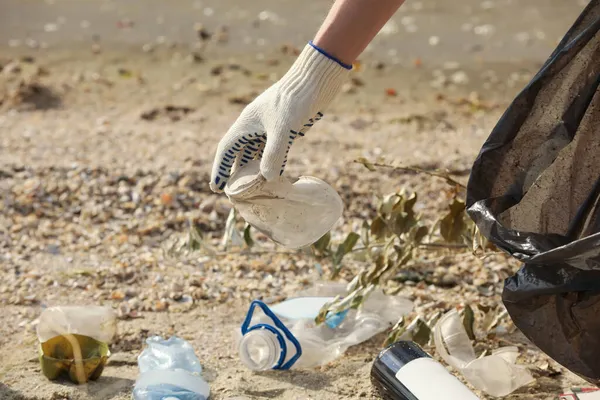 Eldivenli bir kadın çöp torbasıyla sahilde çöp topluyor, yakın plan.