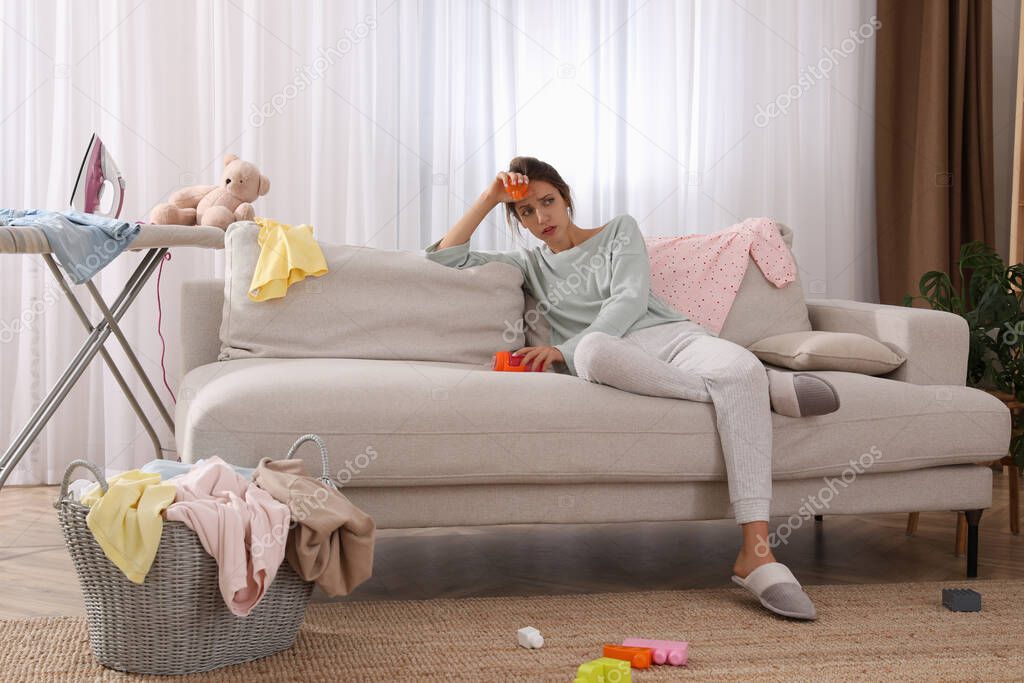Tired young mother sitting on sofa in messy living room