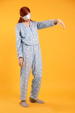 Sarı arka planda uyurgezer halde pijama, maske ve terlik giyen genç bir kadın.