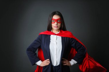 Gri arka planda süper kahraman pelerini ve maskesi takan kendine güvenen bir iş kadını.