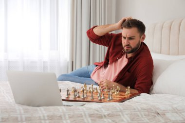 Düşünceli genç bir adam evdeki yatakta online görüntülü sohbet yoluyla partneriyle satranç oynuyor.