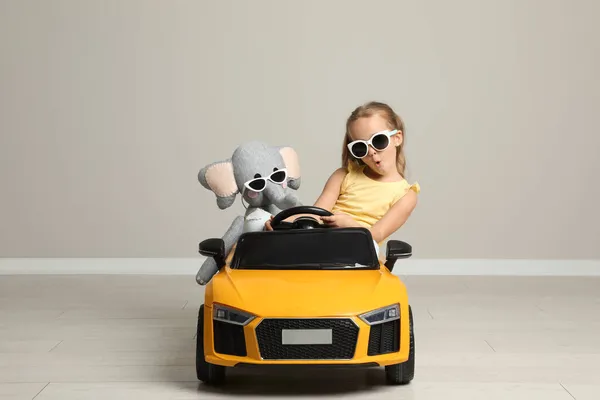 Cute little girl with toy elephant driving children\'s car near grey wall indoors