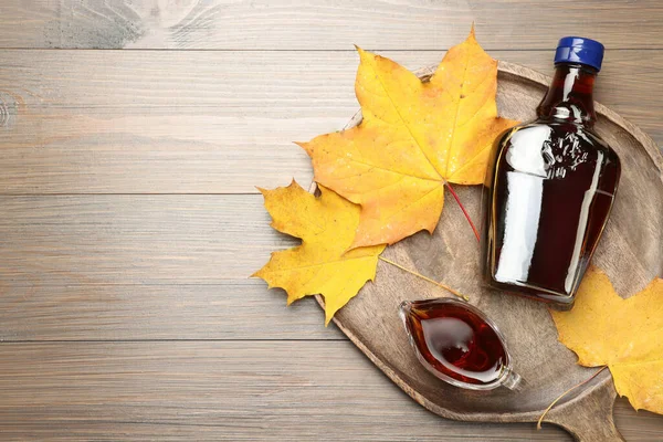 Bottle with tasty maple syrup, gravy boat and dry leaves on wooden table, top view. Space for text