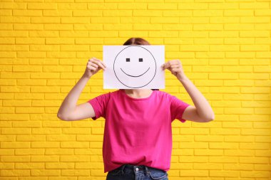 Woman hiding behind sheet of paper with happy face against yellow brick wall clipart