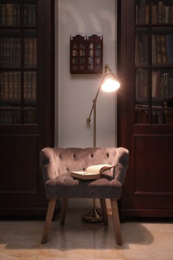 Kütüphanedeki ahşap kitaplıklar arasında kitap ve lambayla rahat bir koltuk.