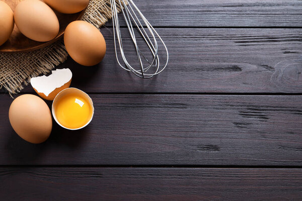 Raw chicken eggs, shell with yolk and whisk on dark wooden table, flat lay. Space for text