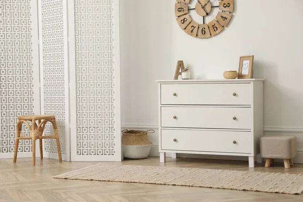 Stylish room with wooden folding screen and chest of drawers near white wall. Interior design