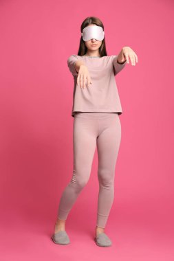 Pembe arka planda uyurgezer halde pijama, maske ve terlik giyen genç bir kadın.