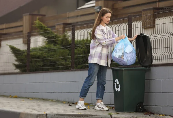 Woman putting garbage bag into recycling bin outdoors