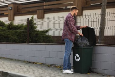 Adam çöp torbasını geri dönüşüm kutusuna koyuyor.