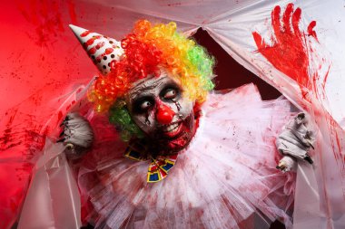 Korkunç palyaço yırtık kanlı plastik filmdeki delikten bakıyor. Cadılar Bayramı partisi kostümü