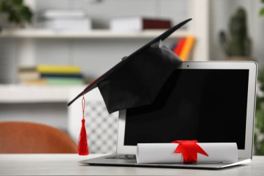 Mezuniyet şapkası, öğrenci diploması ve laptopu beyaz masa içinde.