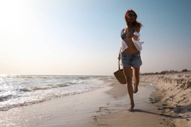 Kumsal çantalı bir kadın güneşli deniz kıyısında yürüyor.