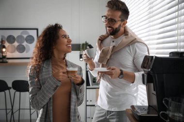 İş arkadaşları sıcak içeceklerle modern kahve makinesinin yanında konuşuyorlar.