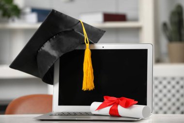 Mezuniyet şapkası, öğrenci diploması ve laptopu beyaz masa içinde.