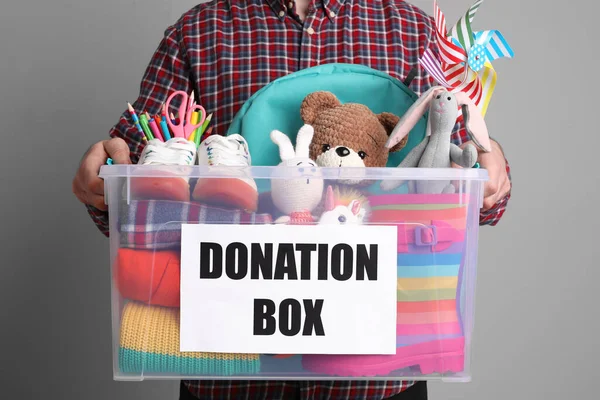 男人拿着装有不同玩具 衣服和文具的捐款箱 灰色背景 — 图库照片