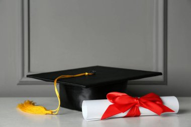 Mezuniyet şapkası ve diploması gri duvarın yanındaki beyaz masada.