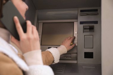 Dışarıda para çekmek için para makinesini kullanırken cep telefonuyla konuşan genç bir kadın.