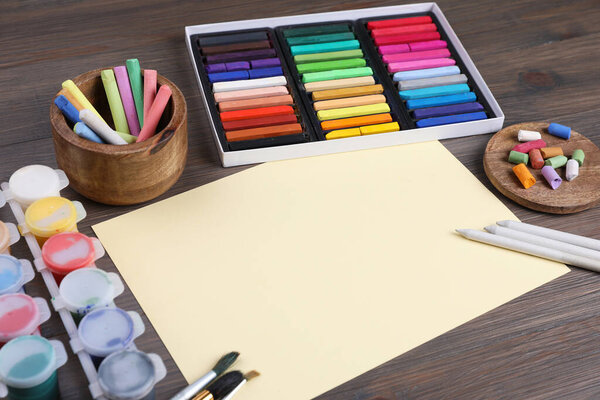 Blank sheet of paper, colorful chalk pastels and other drawing tools on wooden table. Modern artist's workplace