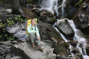 Sırt çantalı mutlu turist dağ şelalesinin yanında dinleniyor.