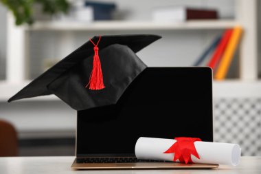 Mezuniyet şapkası, öğrenci diploması ve laptopu beyaz masa içinde.