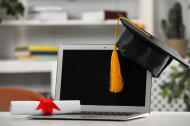Mezuniyet şapkası, öğrenci diploması ve laptopu beyaz masa içinde.