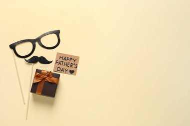 Card with phrase Happy Father's Day, paper glasses, mustache and gift box on pale yellow background, flat lay. Space for text