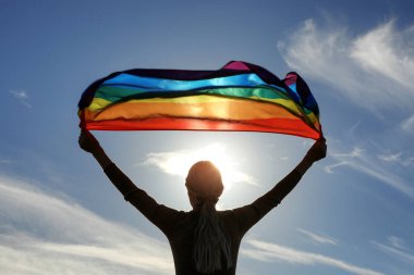 Woman holding bright LGBT flag against blue sky on sunny day, back view