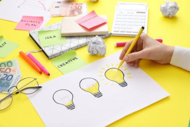 Woman drawing lamp on paper at yellow table, closeup. Startup concept