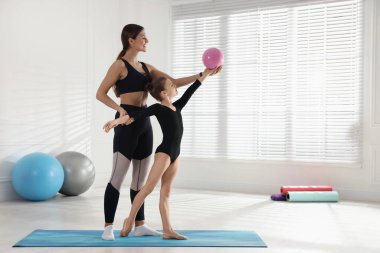 Gymnastic coach helping little girl to do exercise with ball indoors